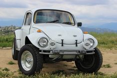 an old vw bug is parked on the side of a dirt road with mountains in the background