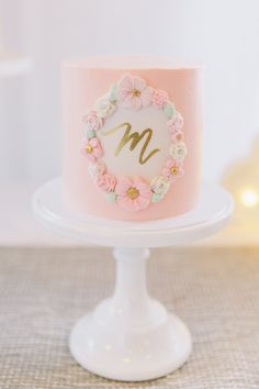 a pink and white cake with flowers around the letter m on a plate in front of a christmas tree