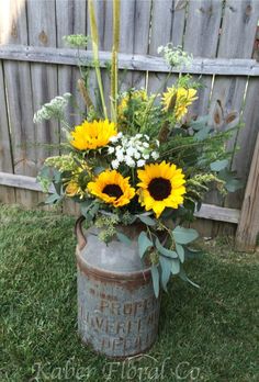 sunflowers and other flowers are in an old milk can on the grass near a fence