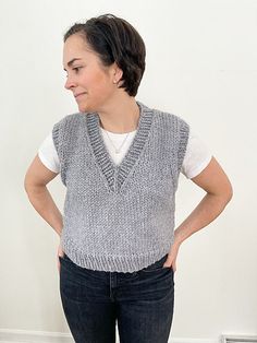 a woman standing in front of a white wall wearing a gray knitted sweater vest