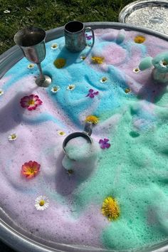 an outdoor table covered in colored sand with flowers on it and two silver mugs