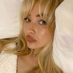 a beautiful blonde woman laying in bed under a white blanket and looking at the camera