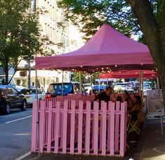 people are sitting under a pink tent on the side of the road in front of some parked cars