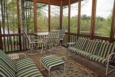 a screened porch with striped furniture and tables on the deck overlooking a lake in the distance