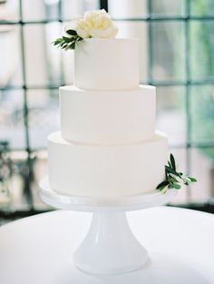 a white wedding cake sitting on top of a table next to a large window and greenery