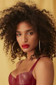 a woman with curly hair wearing a red dress and hoop earrings, posing in front of a yellow background