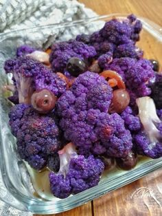 purple cauliflower is in a glass dish on a wooden table with other vegetables