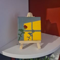 a small easel with a flower painted on it sitting on a shelf in front of a window