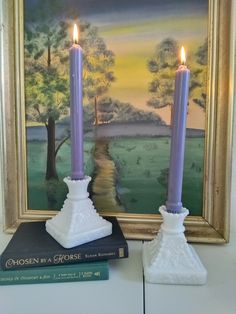 two candles are sitting next to a book on a table in front of a painting