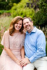 a man and woman are sitting on a bench in front of some tall grass, smiling at the camera