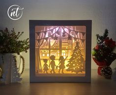 a paper cut christmas scene with two children in front of a lit up tree, surrounded by flowers and pine cones