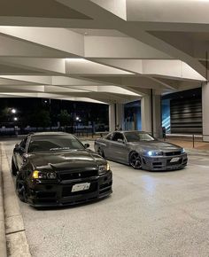 two cars are parked in an empty parking lot at night, one is black and the other is silver