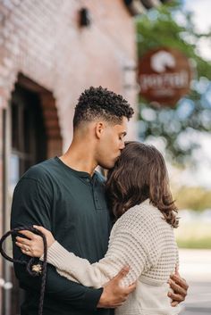 a man and woman standing next to each other in front of a brick building with their arms around each other