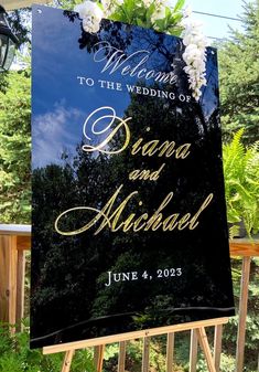 a welcome sign for a wedding with flowers on the front and back of it, hanging from a wooden fence