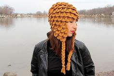 a woman wearing a crocheted hat standing next to the water with her hands in her pockets