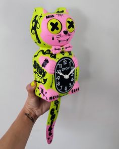 a pink and yellow cat clock is held up by a person's hand with tattoos on it