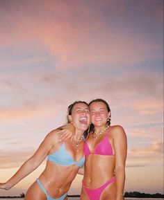 two women in bikinis standing on the beach at sunset with their arms around each other