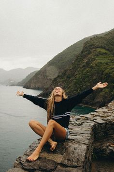 a woman sitting on the edge of a cliff with her arms outstretched in front of water