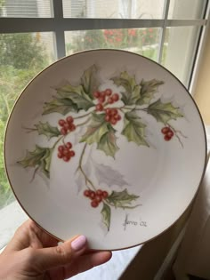 a hand holding a white plate with holly and red berries on it, in front of a window