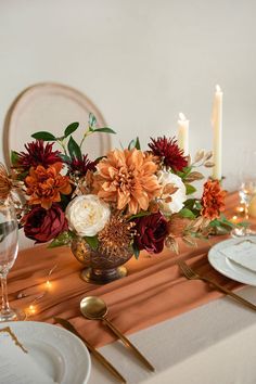 an arrangement of flowers in a vase on a dining room table with candles and place settings