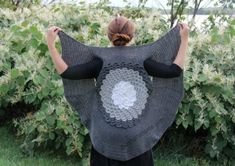 a woman is standing in front of bushes with her back to the camera, holding up a gray crocheted shawl