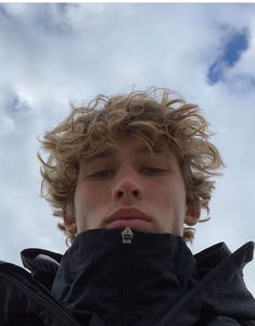 a young man with curly hair standing in front of the sky looking up at something