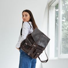 a woman carrying a brown leather backpack in front of a white wall and window with her hands on her hips