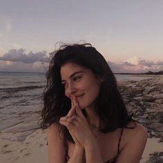 a woman sitting on top of a sandy beach next to the ocean with her finger in her mouth