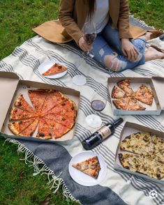 a woman sitting on the grass with four pizzas in front of her and two bottles of wine