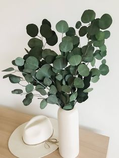 a white vase filled with green leaves next to a hat on top of a wooden table