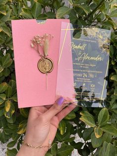 a person holding up two pink and blue wedding cards in front of some greenery