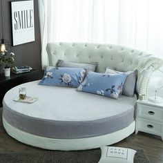 a white round bed with blue and grey pillows on it in a room next to a window