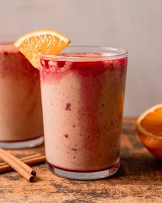 two glasses filled with smoothie next to an orange slice and cinnamon sticks on a table