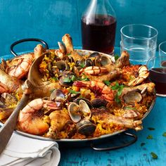 a large pan filled with seafood and rice next to two glasses on a blue table