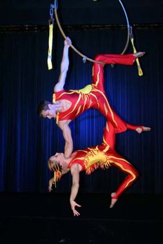 two men performing aerial acrobatic tricks on a circus ring, one in red and the other in yellow