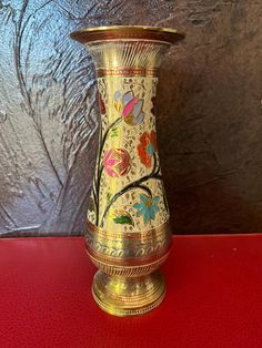 a gold vase with colorful flowers painted on the side sitting on a red table top