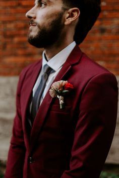 a man in a red suit and tie standing next to a brick wall wearing a flower boutonniere