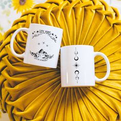 two white coffee mugs sitting next to each other on a yellow pillow with sunflowers in the background