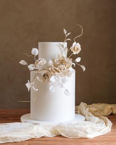 a white wedding cake decorated with flowers and greenery on a wooden table next to a cloth