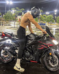 a shirtless man standing next to a black motorcycle in the rain at night with its lights on