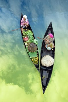 two boats with people in them floating on the water, one is filled with plants