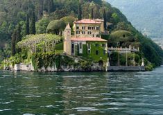 an old house on top of a hill next to the water with trees and bushes growing all around it