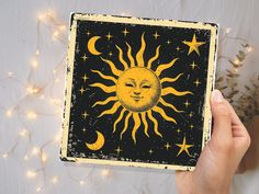 a hand holding up a small square painting with the sun and moon on it, surrounded by string lights