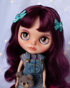 a close up of a doll with purple hair and blue overalls holding a teddy bear