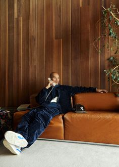 a man sitting on top of a brown couch talking on a cell phone next to a potted plant