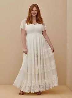 a woman wearing a white dress and sandals standing in front of a beige wall with her hands on her hips