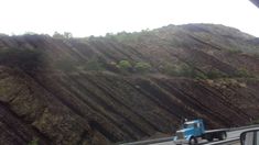 a blue truck driving down a road next to a tall mountain covered in green trees