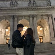 a man and woman standing in front of a building with columns on each side, looking at each other