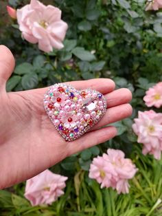 a hand holding a pink heart shaped ring in front of some flowers and greenery