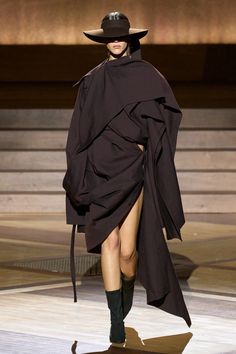 a woman in a black dress and hat walks down the runway with her coat draped over her shoulders